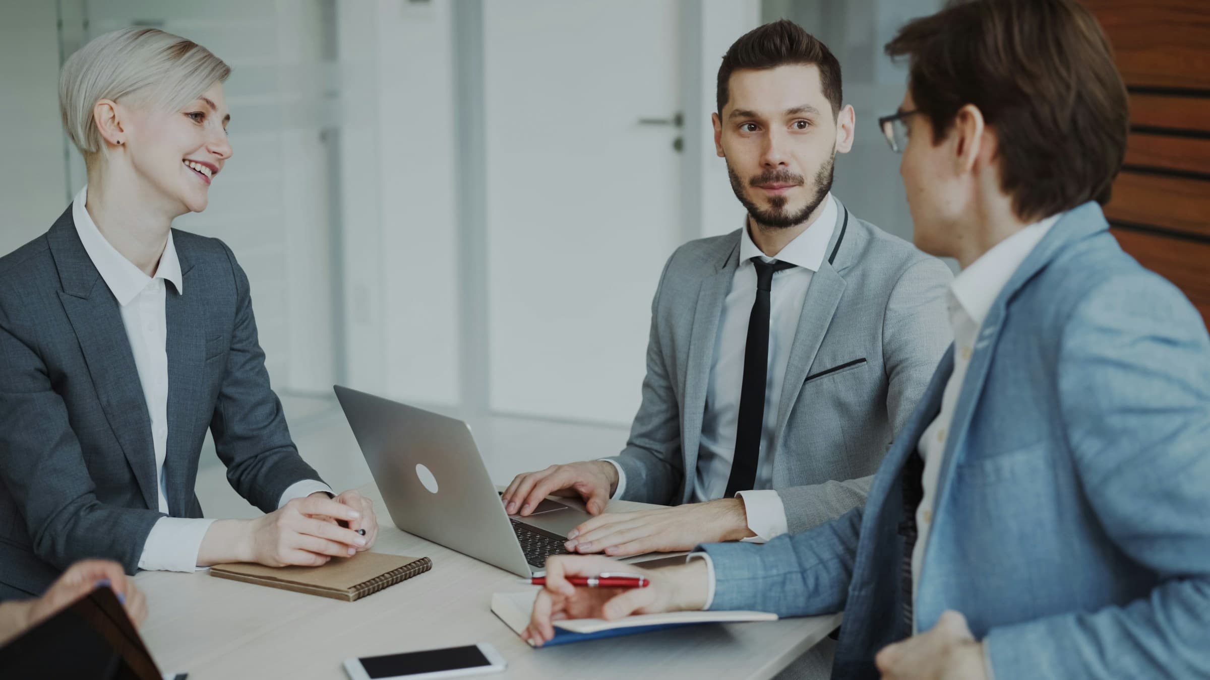 A hiring team of three colleagues discussing a role brief together at a shared table, one typing on a laptop.