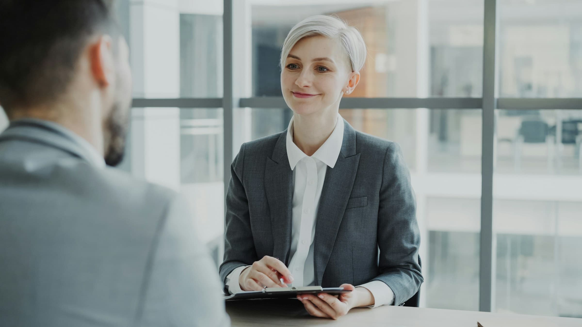 A recruiter in a grey blazer interviews a candidate across a table, holding a clipboard and pen.