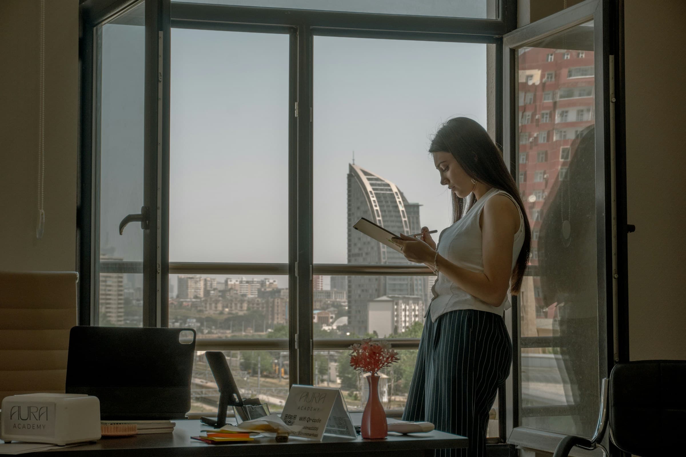 A woman reading a document by a tall window in a quiet office.