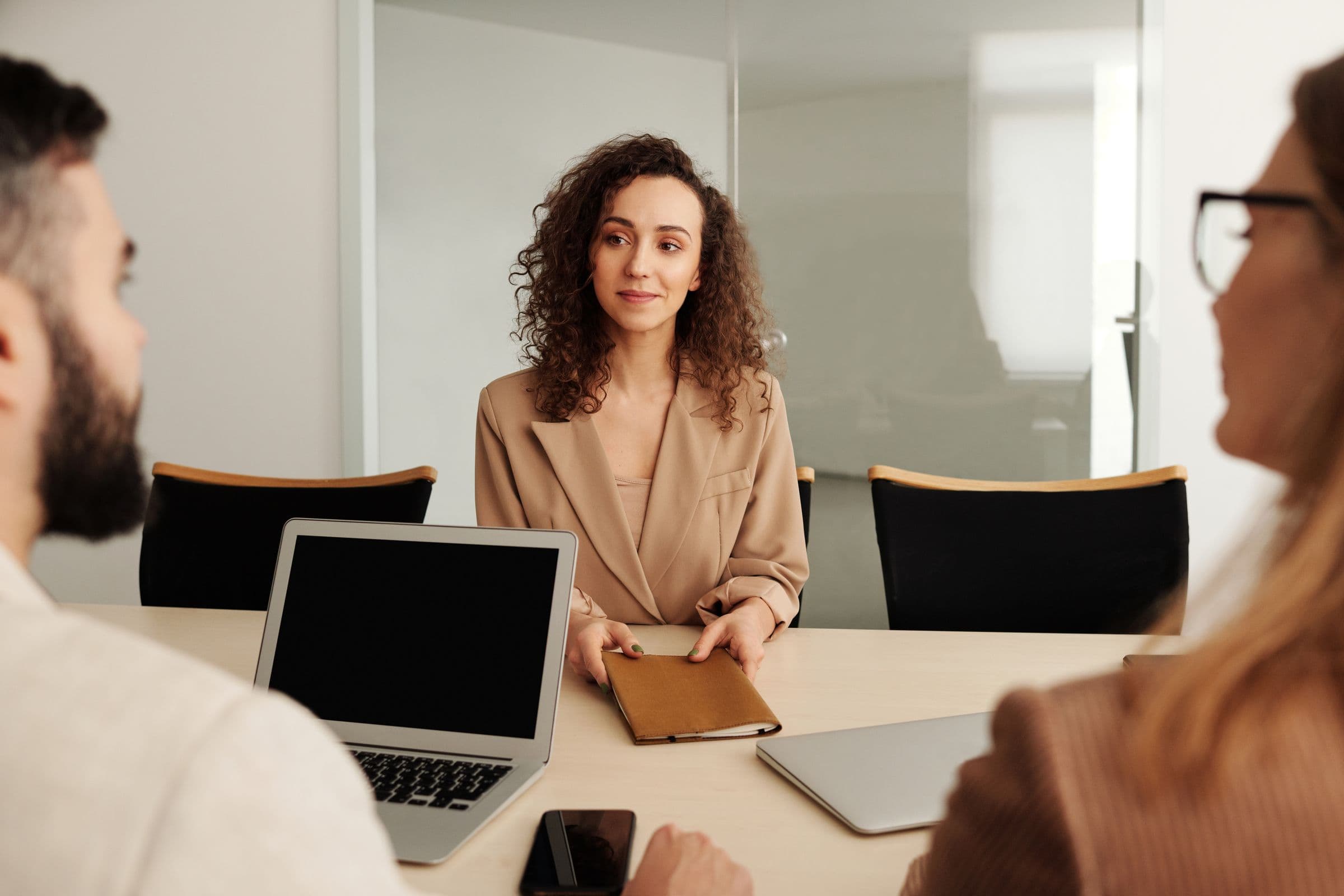 A candidate at ease across the table from two interviewers, handing over a notebook in a sunlit meeting room.