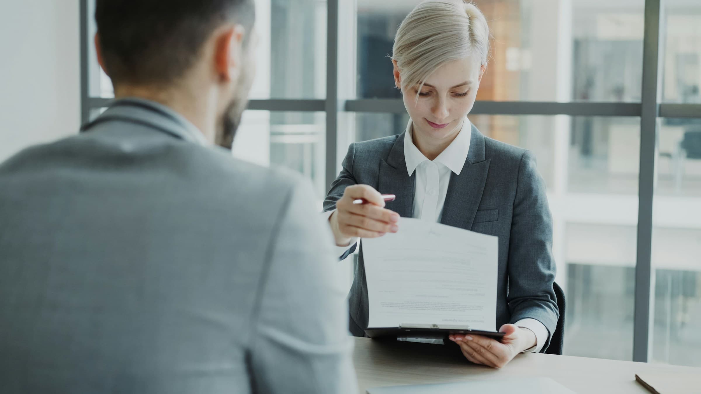 A recruiter hands a hiring manager a candidate profile on a clipboard across the table.