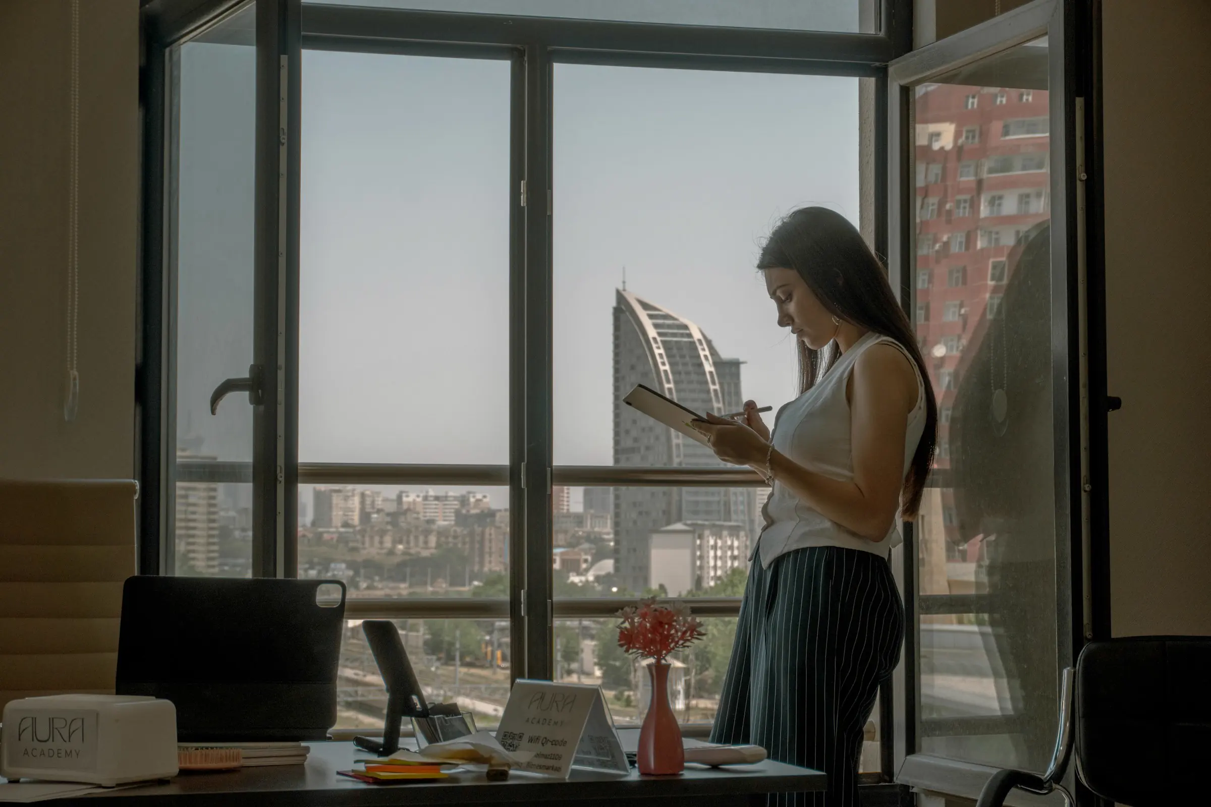 A woman writing on a notepad while standing at a tall office window, a city skyline behind her.