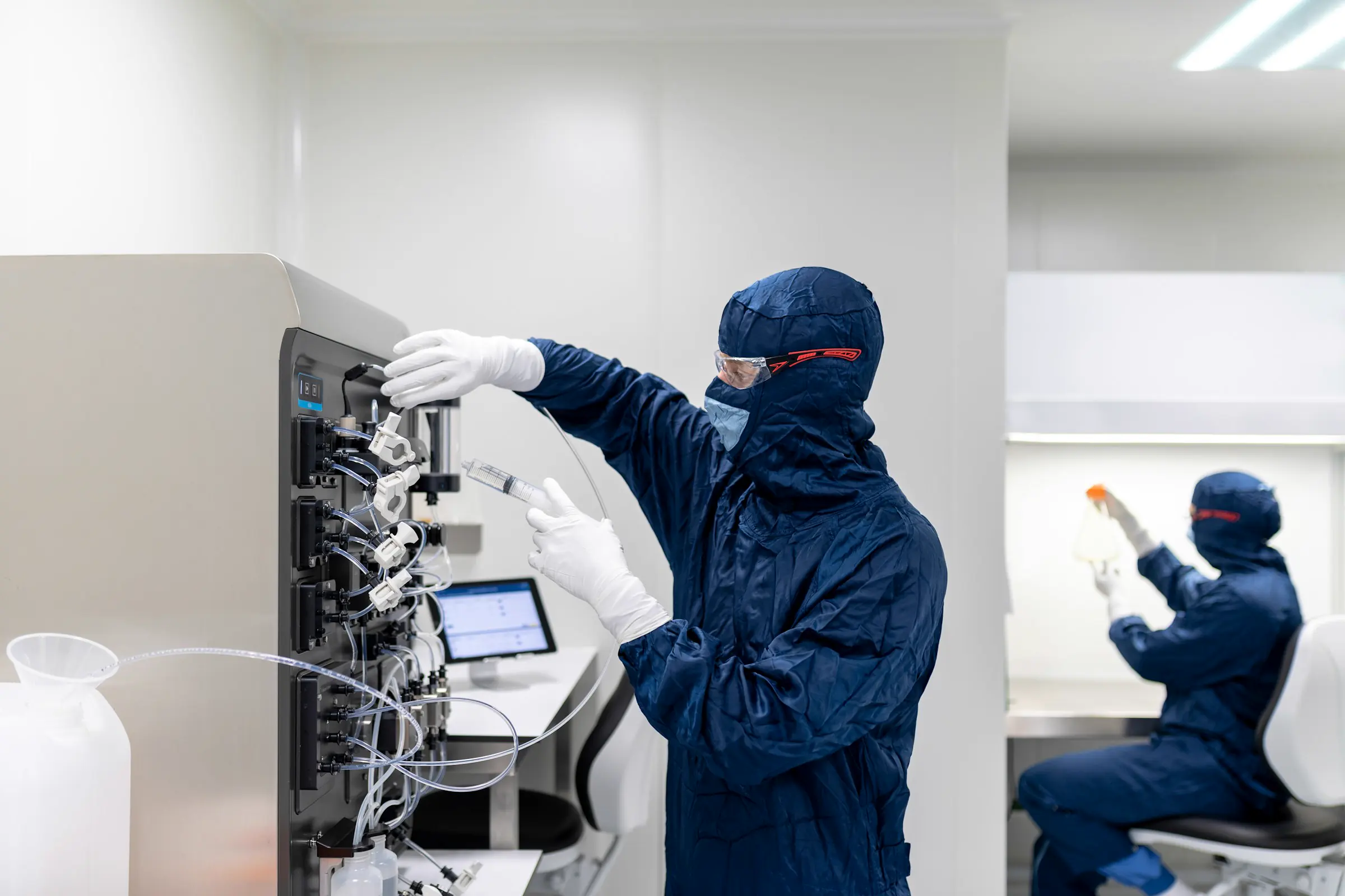A technician in a full cleanroom gown and goggles setting up tubing on a filling machine, with a colleague working in the background.