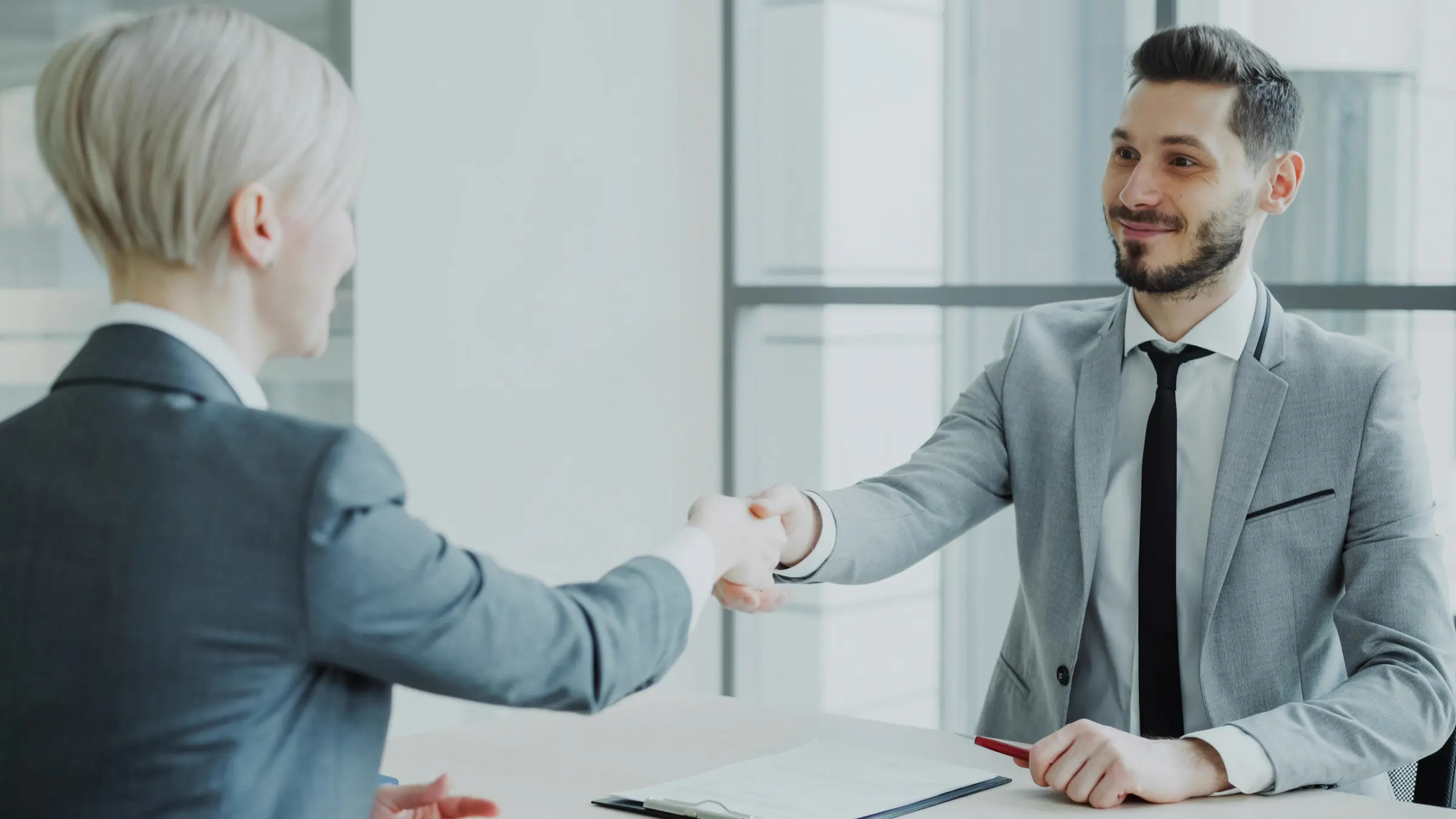 A hiring manager and a new hire shake hands across the table, both smiling.